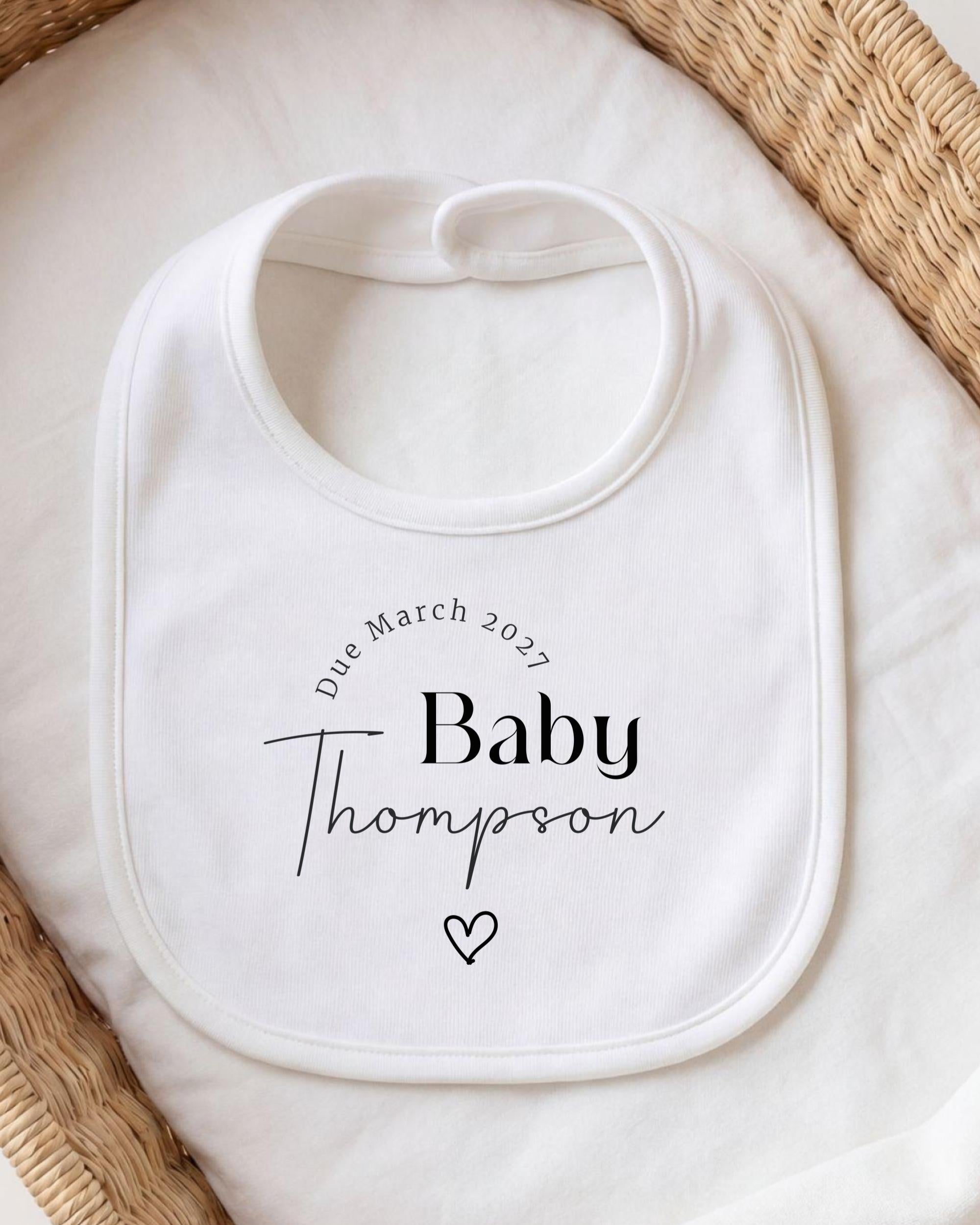 A white bib with personalized text and a heart symbol, placed on a white cloth.