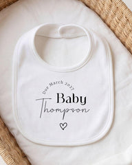 A white bib with personalized text and a heart symbol, placed on a white cloth.
