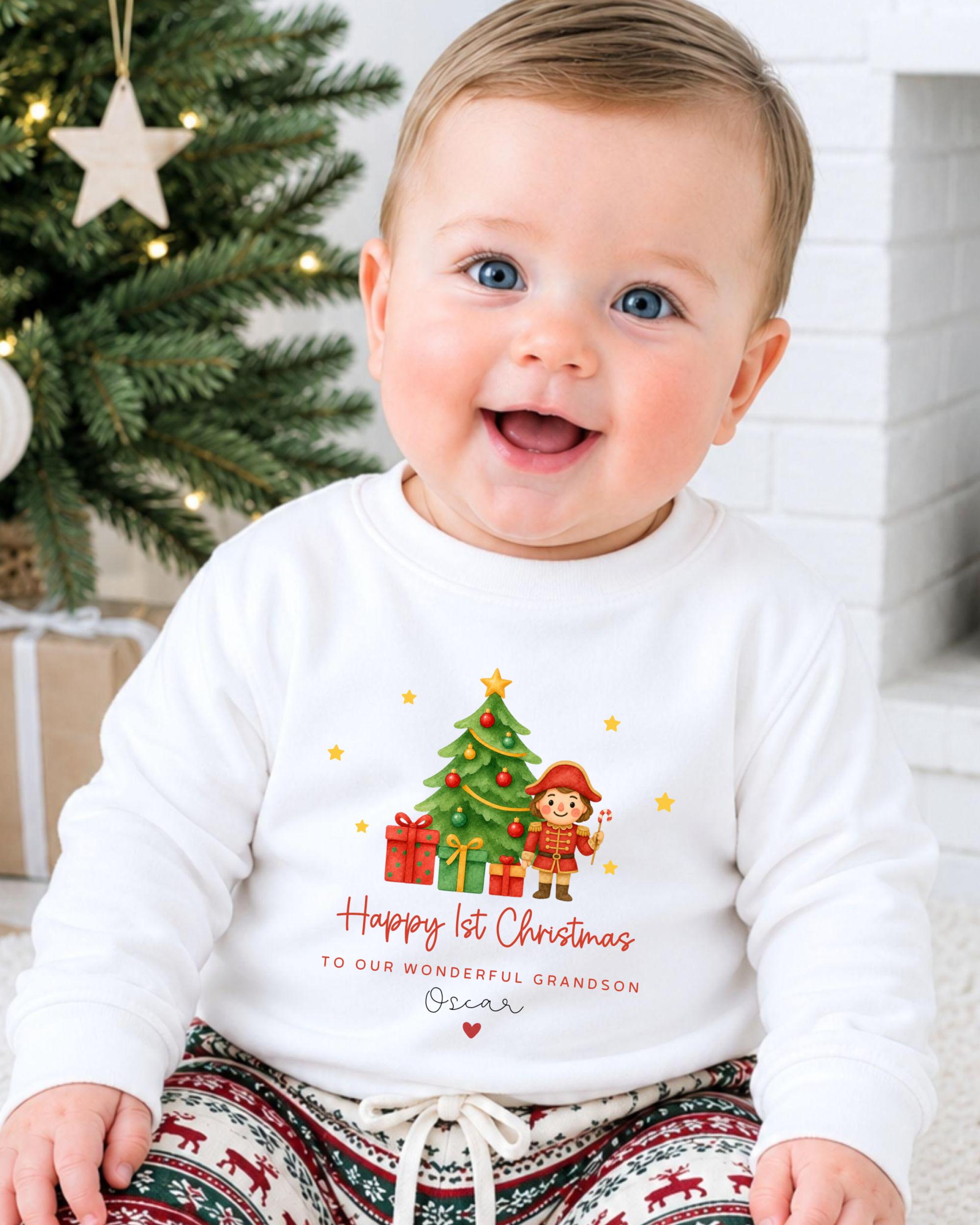 A smiling baby wearing a white sweatshirt with a Christmas tree and presents on it, sitting in front of a Christmas tree.