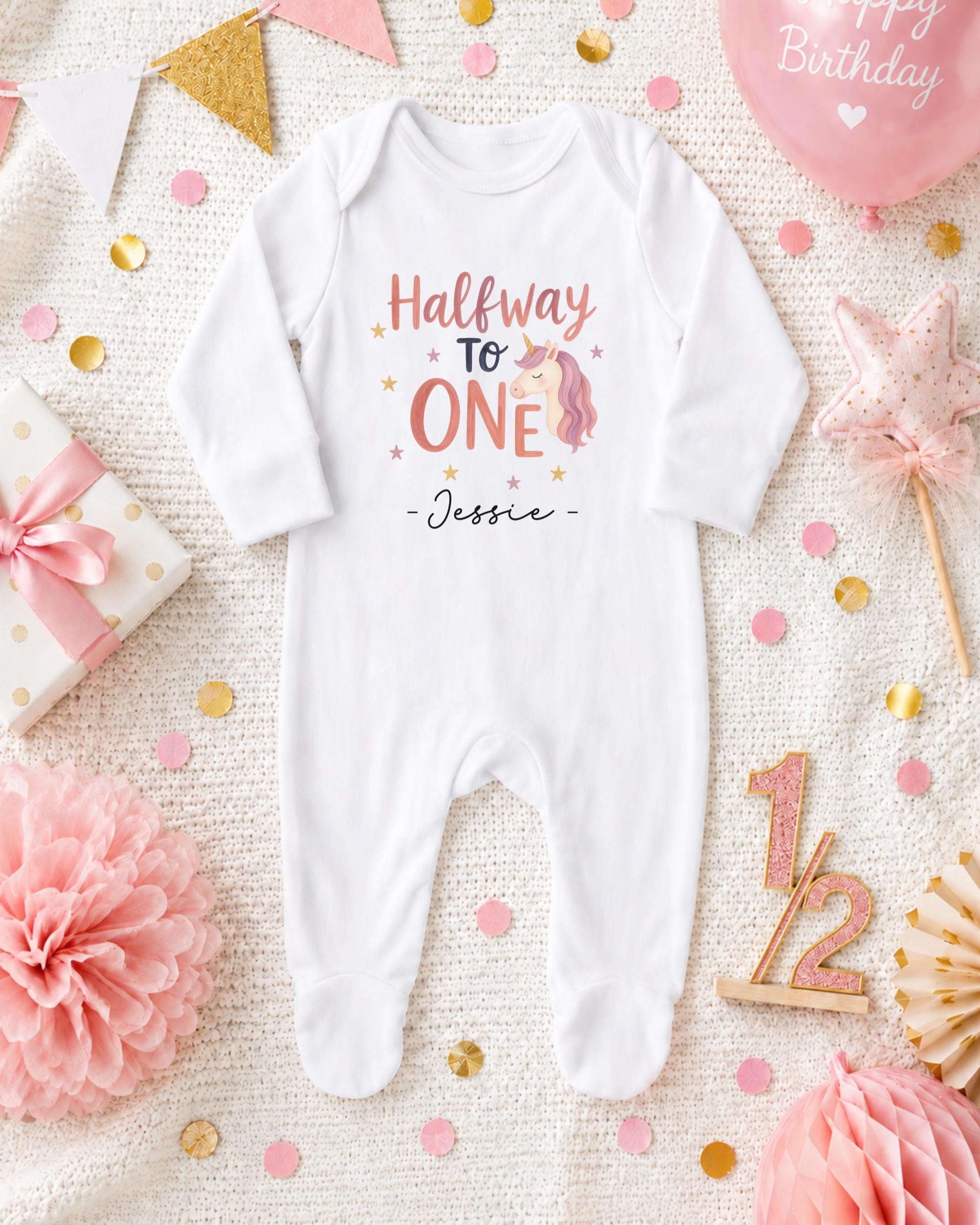 A white onesie with the text "Halfway to one" and a unicorn graphic, surrounded by confetti, a birthday balloon, and other baby-related items.