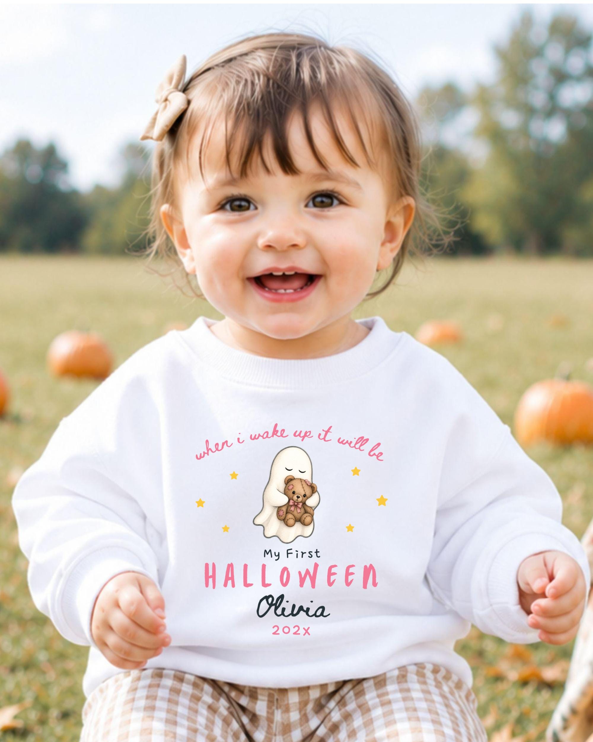 A smiling baby girl wearing a white sweater with a Halloween-themed design, sitting in a field with pumpkins in the background.