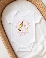 A white onesie with a graphic design on it, featuring a bottle of beer and a bottle of milk, along with the text "Happy Birthday Daddy Lots of Love Annie" printed on it.