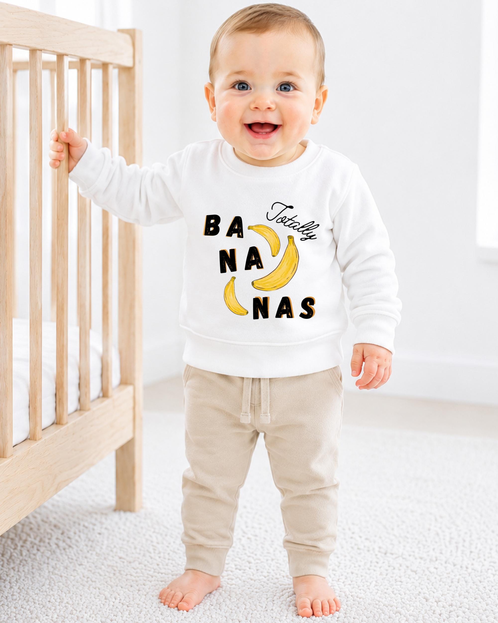 A smiling baby boy wearing a white sweatshirt with a banana graphic and the text "TotALLY BANANA" is standing next to a wooden crib.