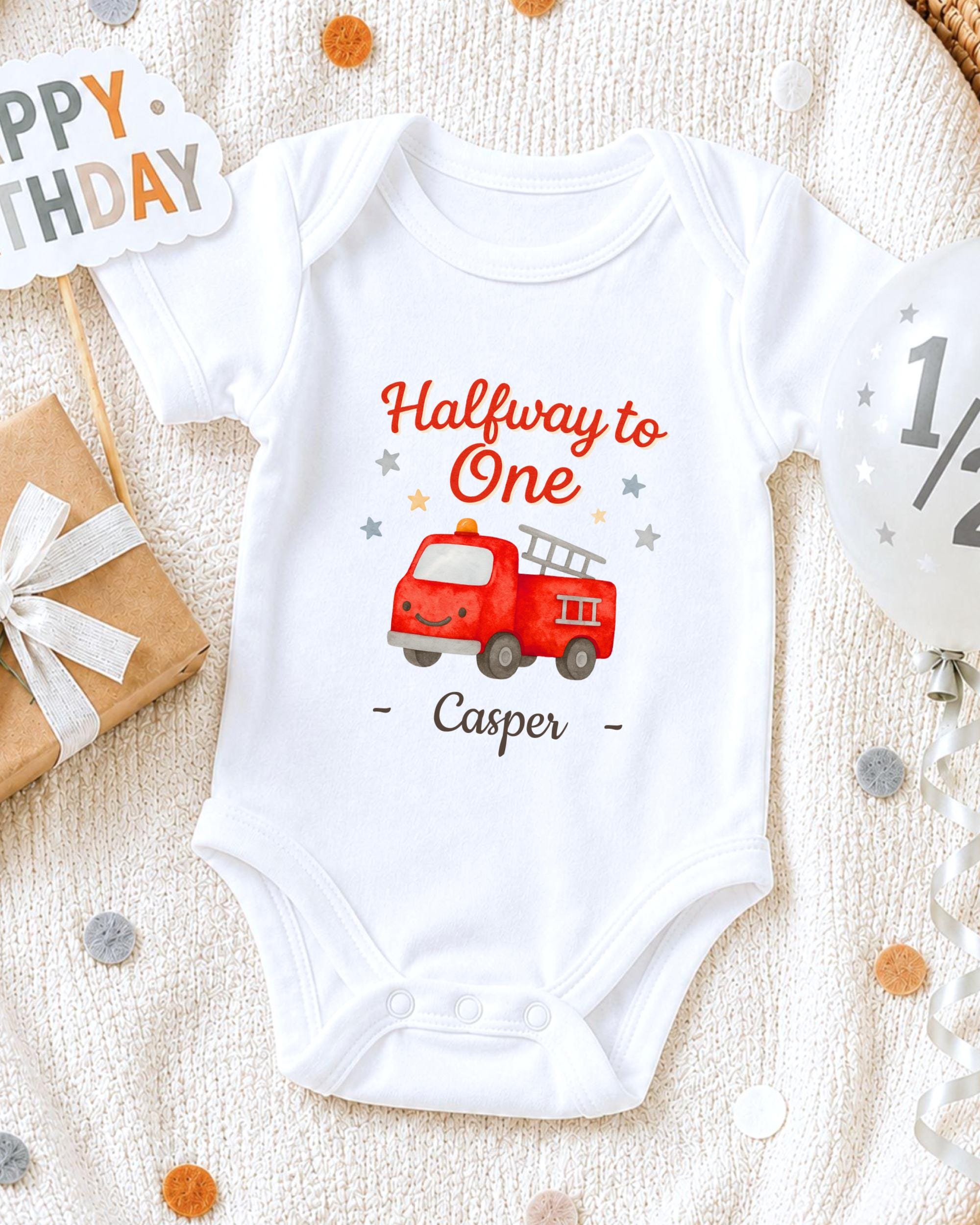 A white onesie with a red fire truck graphic and the text "Halfway to One Casper" printed on it, along with a "Happy Birthday" sign and a gift box.