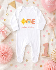 A white onesie with the name "Alexander" printed on it, surrounded by birthday decorations such as a pink star, a pink flower, and confetti.