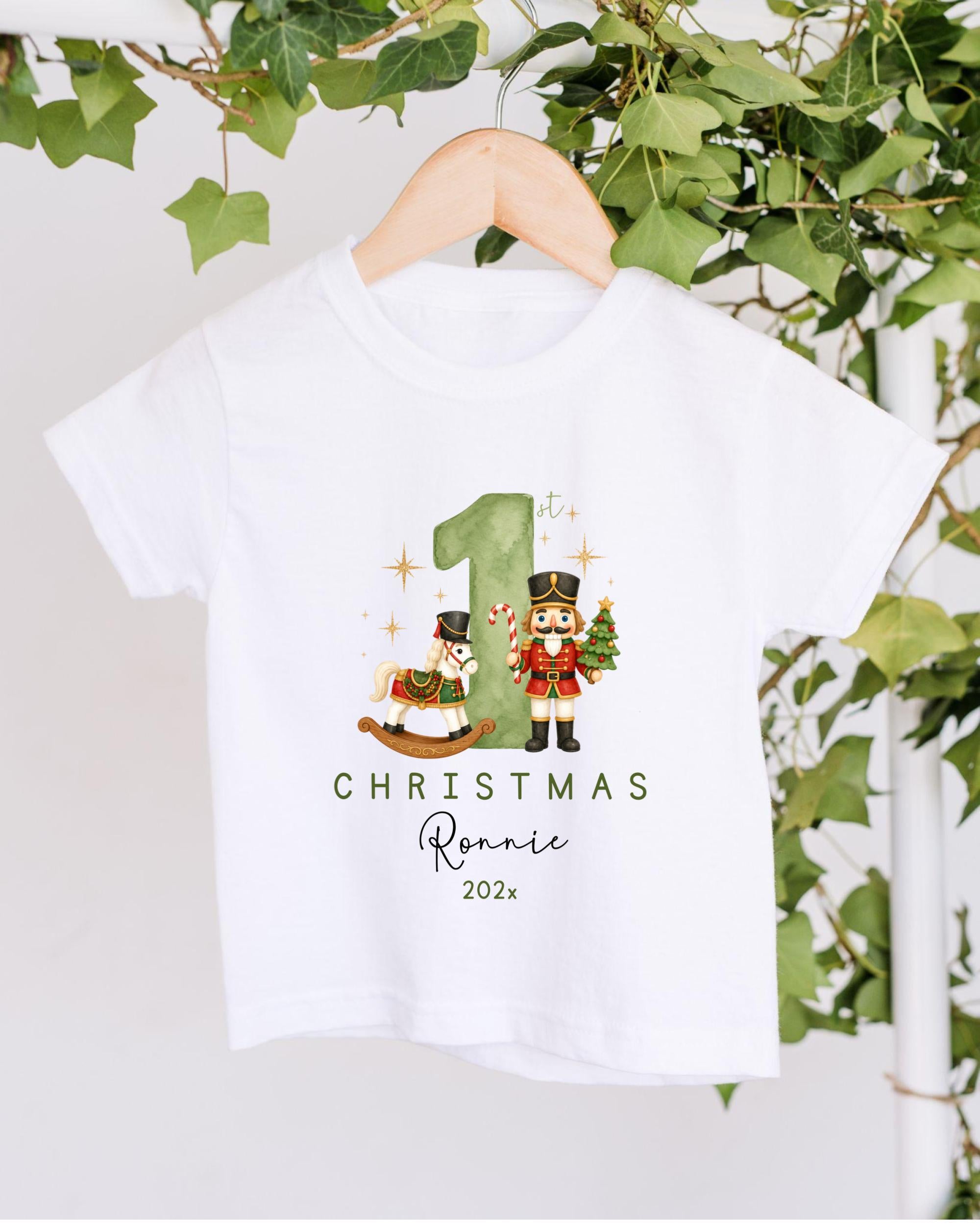 A white t-shirt with a Christmas-themed design hanging on a wooden hanger. The design features a nutcracker character and a horse, along with the text "1st Christmas Ronnie 202x".