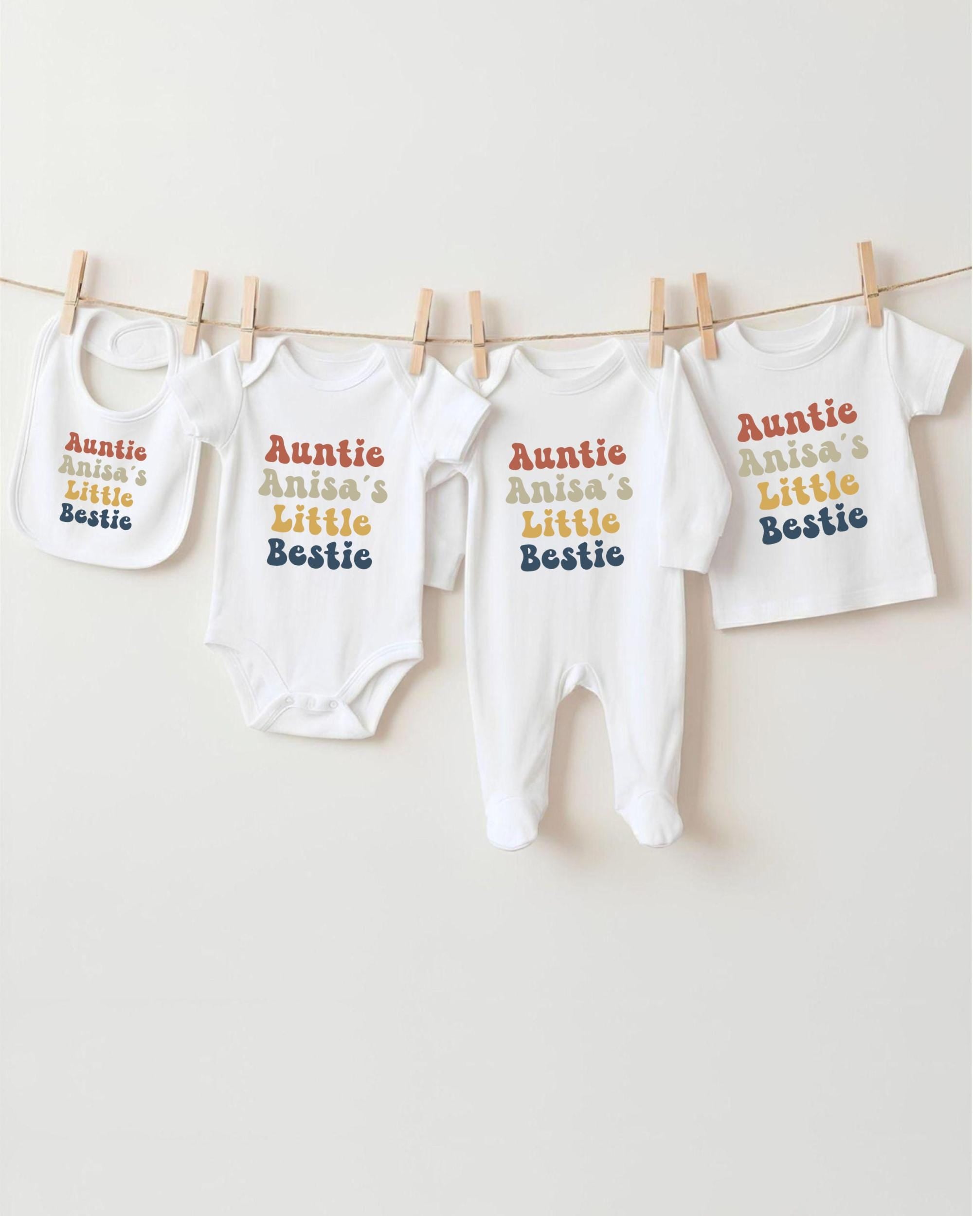 A row of white baby clothes with the text "Auntie Anisa's Little Bestie" printed on them, hanging on a clothesline.