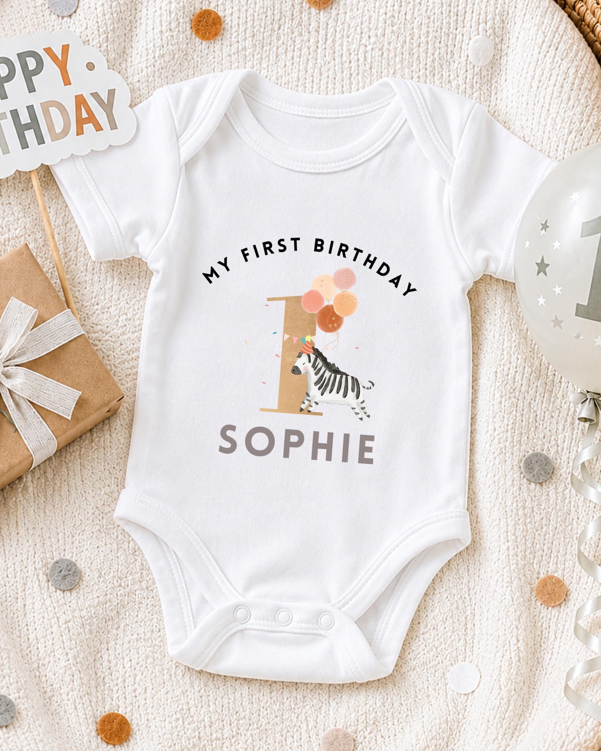 A white onesie with a zebra and the text "My first birthday Sophie" printed on it, along with a "1" and a birthday sign.