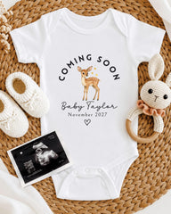 A white onesie with a deer graphic and the text "Coming soon Baby Taylor November 2027" is displayed on a woven placemat, accompanied by a crocheted rabbit toy and a baby shoe.