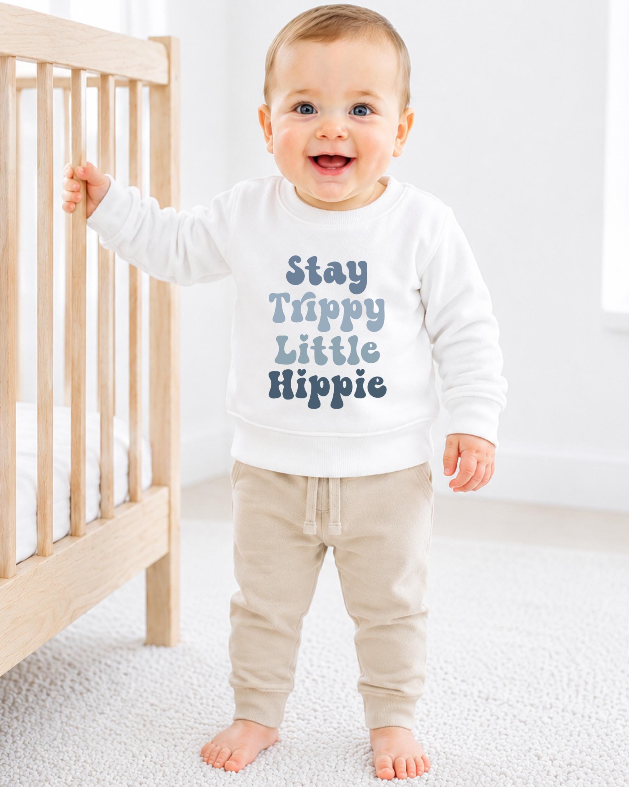A smiling baby boy wearing a white sweatshirt with the text "Stay Trippy Little Hippie" printed on it, standing next to a wooden crib.