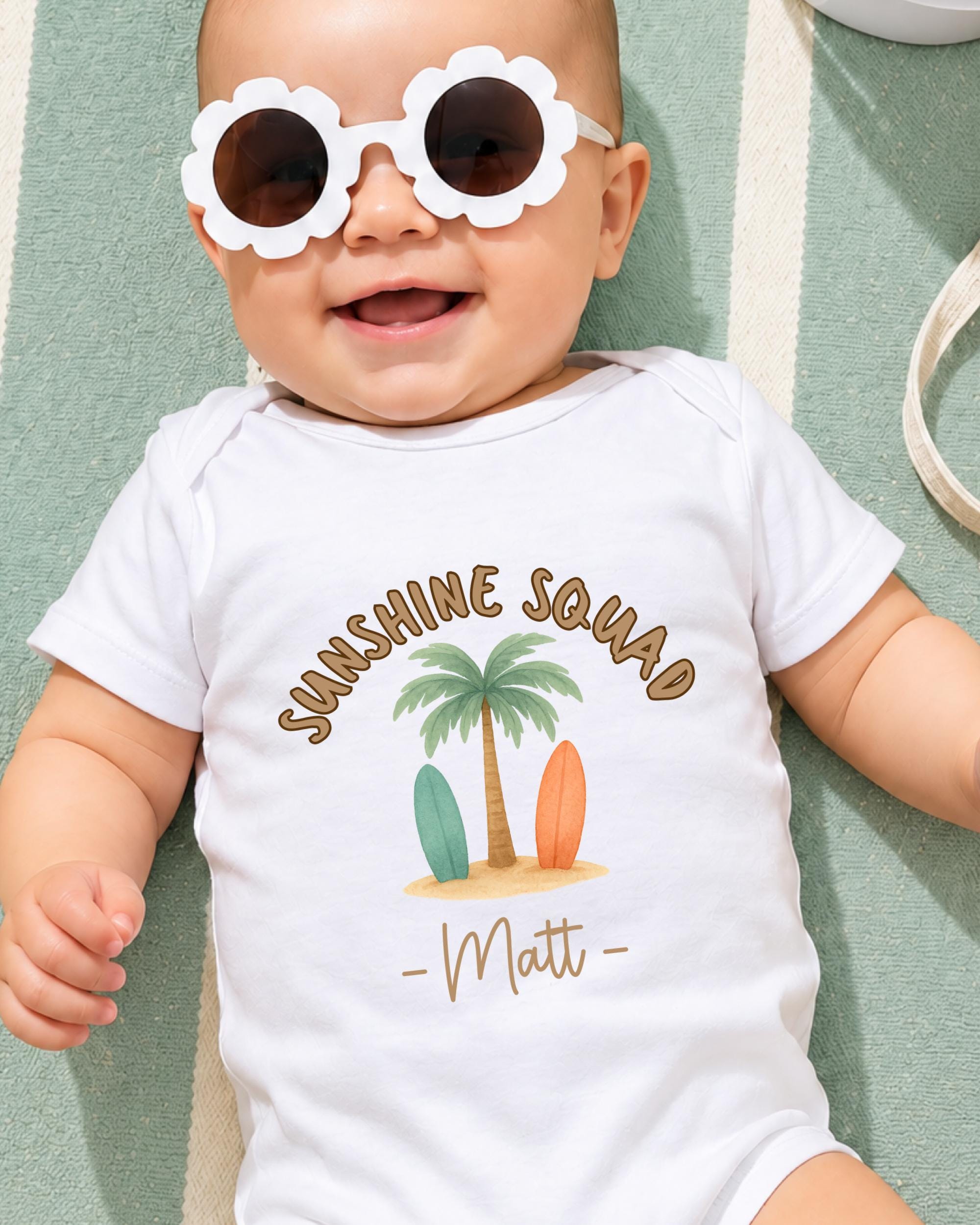 A baby wearing a white onesie with a graphic of a palm tree and surfboards, smiling and wearing sunglasses.