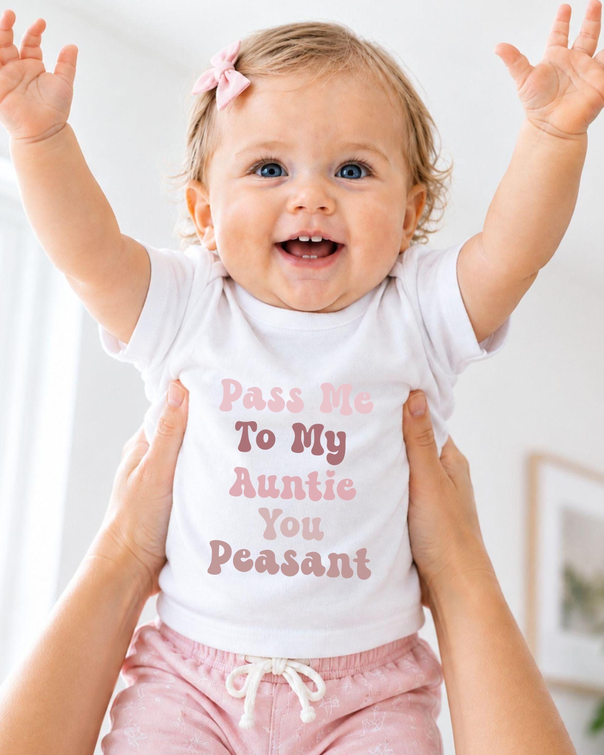 A smiling baby girl wearing a white shirt with text and pink shorts is being held up by someone's arms.