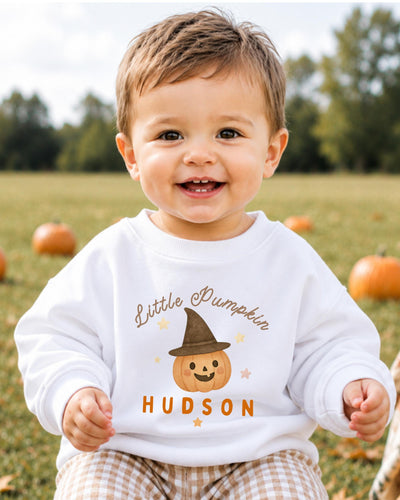 A smiling baby boy wearing a white sweatshirt with a pumpkin and the name "Hudson" printed on it, sitting in a field with pumpkins in the background.