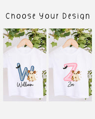 two white t-shirts with personalized letter designs on them. The letters are "W" and "Z" with a cute cartoon character below each letter. The t-shirts are hanging on a wooden hanger, and there are green plants in the background.