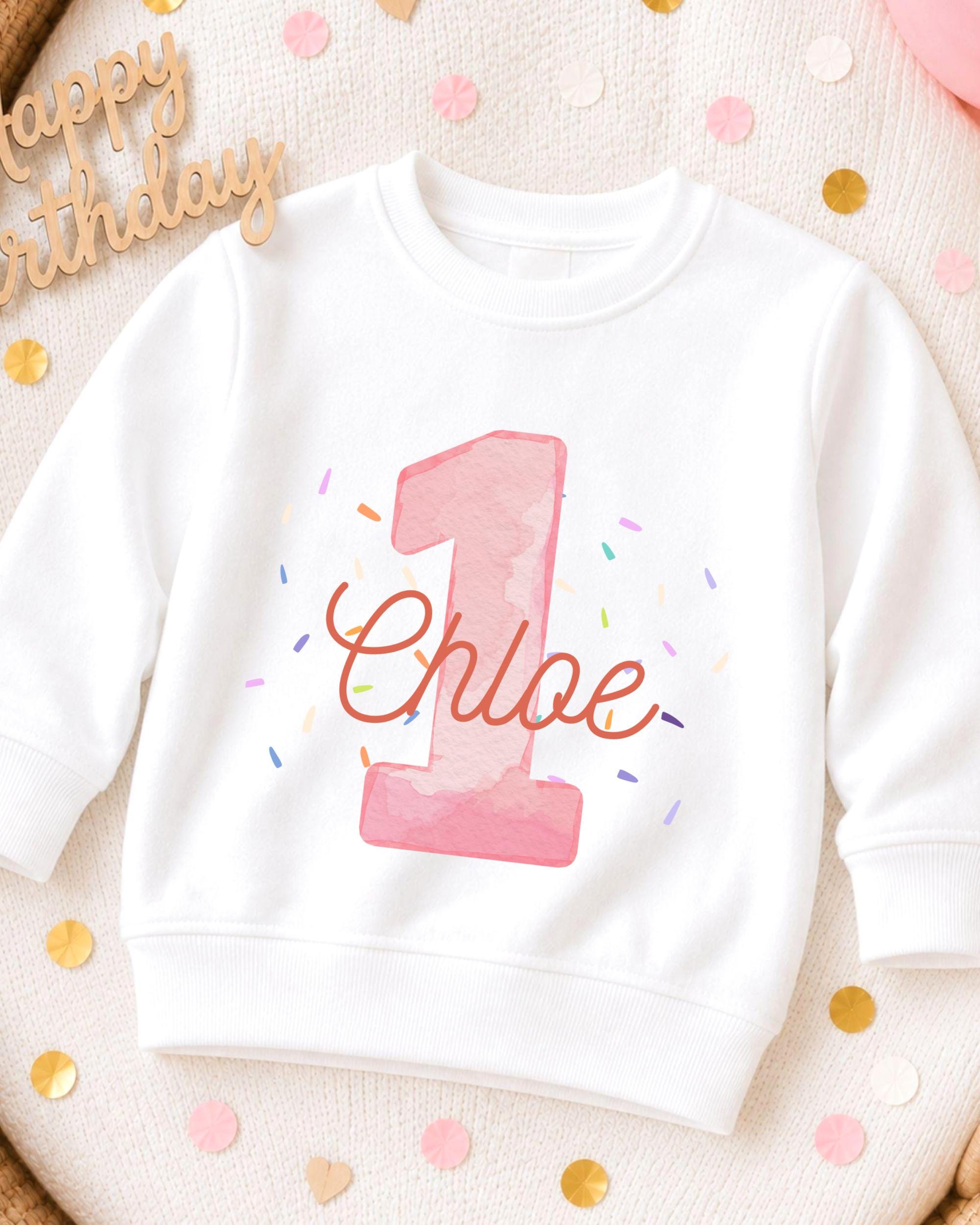 A white sweatshirt with a pink number one and the name "Chloe" printed on it, surrounded by confetti.