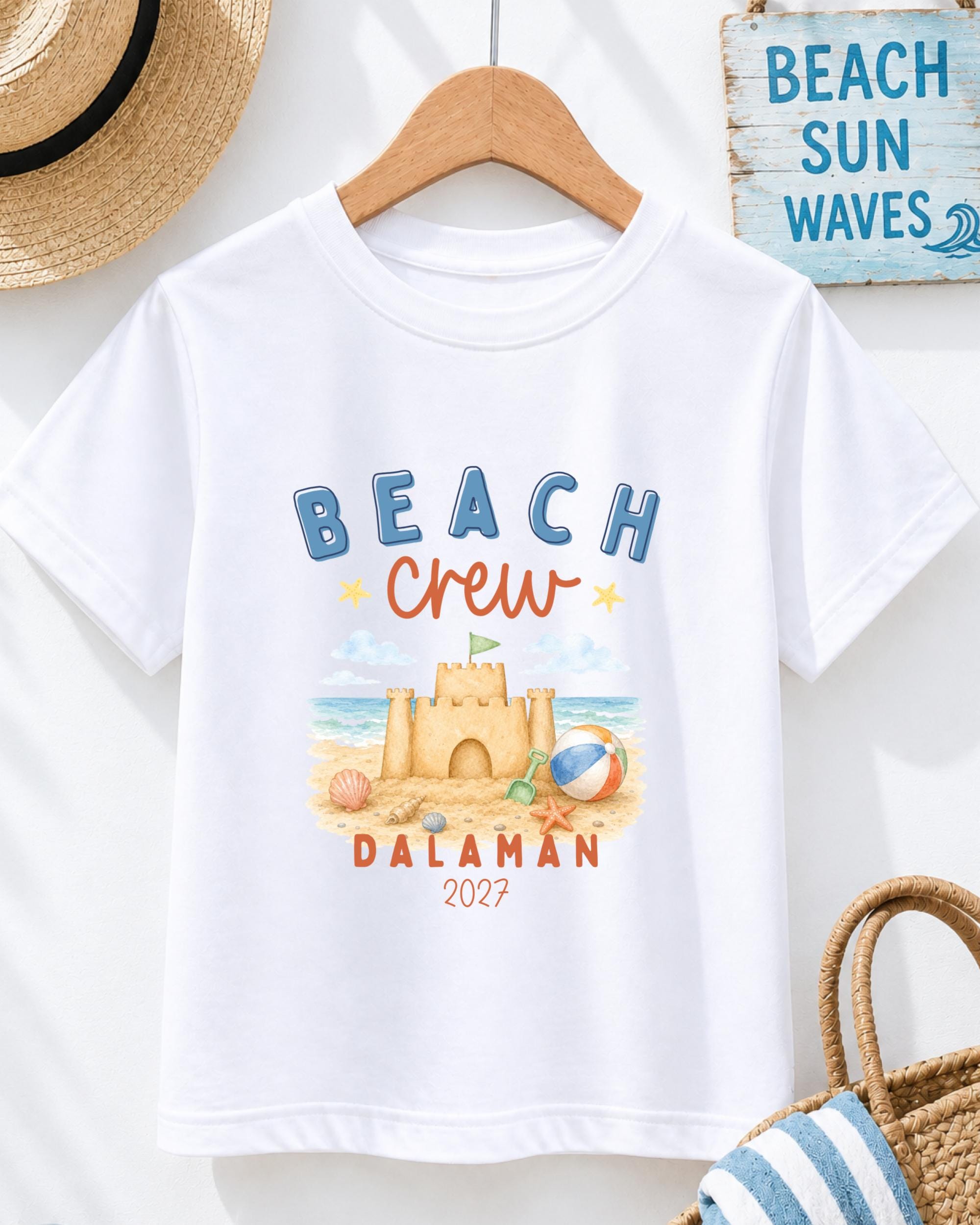 A white t-shirt with a beach-themed design, including a castle, beach ball, and sandcastle, along with the text "BEACH CREW DALAMAN 2027".