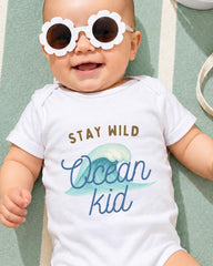 A smiling baby wearing a white onesie with the text "STAY WILD Ocean Kid" printed on it, lying on a green and white striped surface.