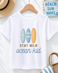 A white t-shirt with a graphic design of three surfboards and the text "STAY WILD ocean kid" printed on it, along with a wooden hanger and a straw hat.