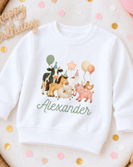 A white sweatshirt with a cartoon illustration of farm animals, including a cow, pig, and sheep, along with balloons and a birthday message.