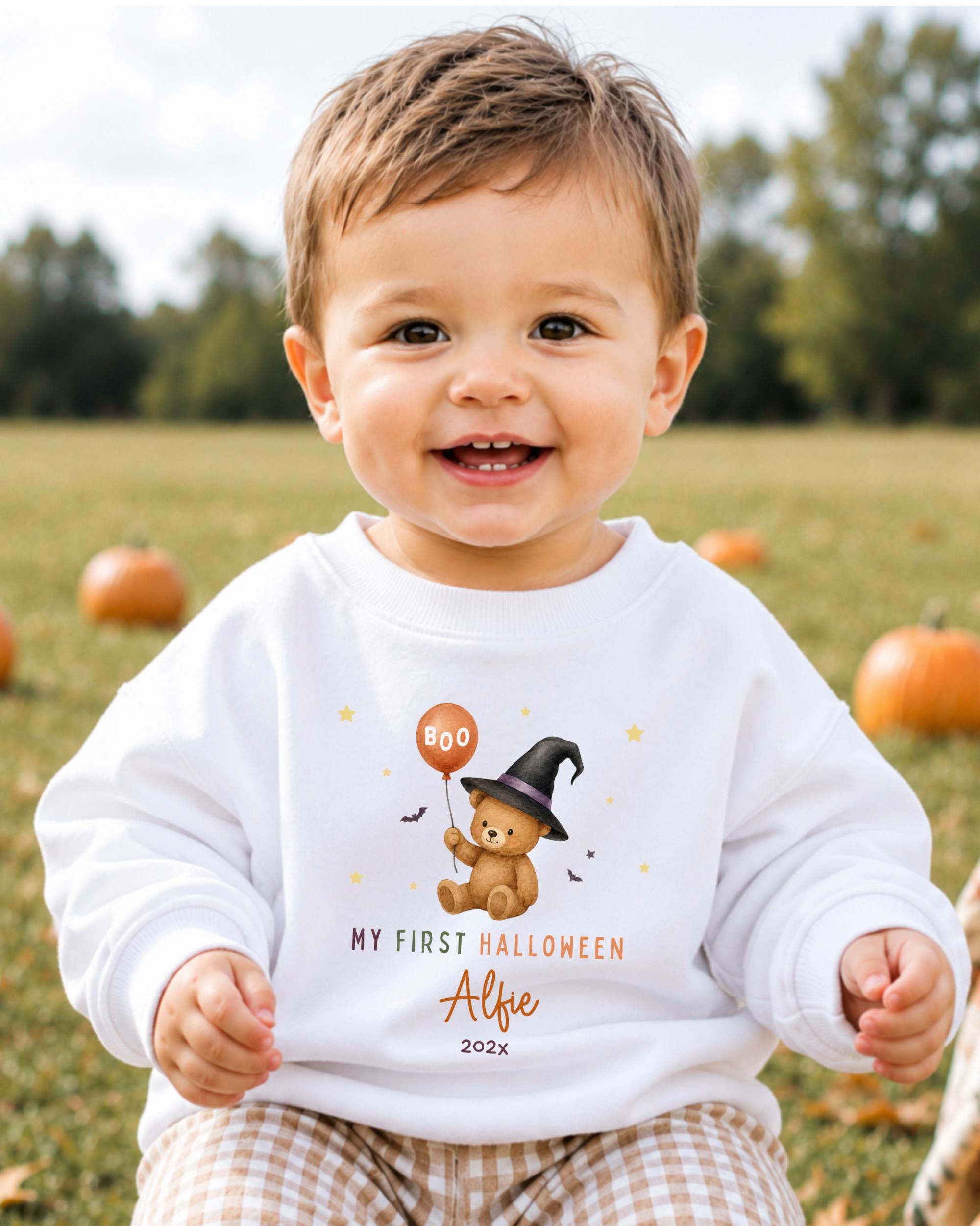 A smiling baby boy wearing a white sweatshirt with a cartoon teddy bear and Halloween-themed text.