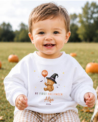 A smiling baby boy wearing a white sweatshirt with a cartoon teddy bear and Halloween-themed text.