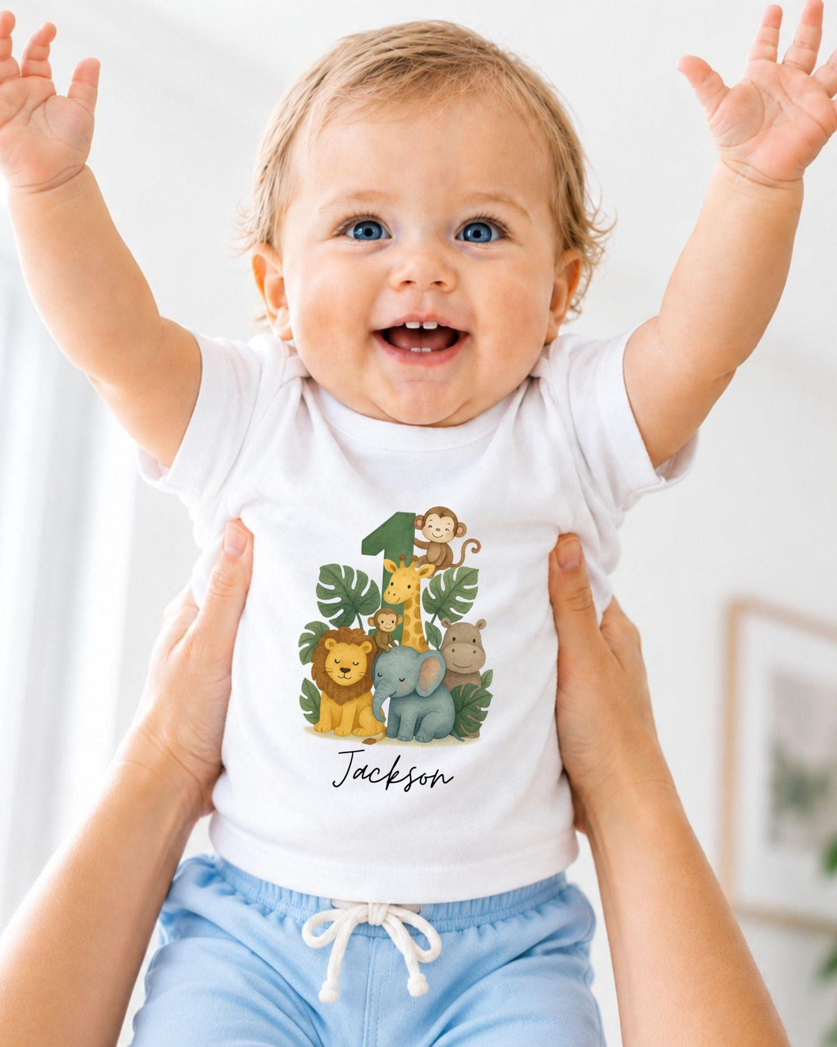 A smiling baby wearing a white t-shirt with a jungle-themed graphic and the name "Jackson" printed on it, being held up by someone's arms.