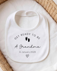 A white bib with the text "GET READY TO BE A GRANDMA In January 2028" printed on it, along with a small footprint symbol.