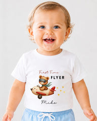 A smiling baby wearing a white t-shirt with a cartoon image of a bear piloting a plane and the text "First Time Flyer Mike" printed on it.