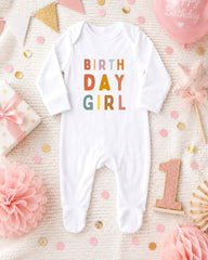 A white onesie with the text "BIRTH DAY GIRL" printed on it, surrounded by birthday decorations such as confetti, a balloon, and a pink star.