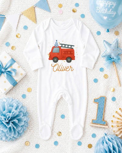 A white onesie with a red firetruck design, surrounded by birthday decorations and confetti.