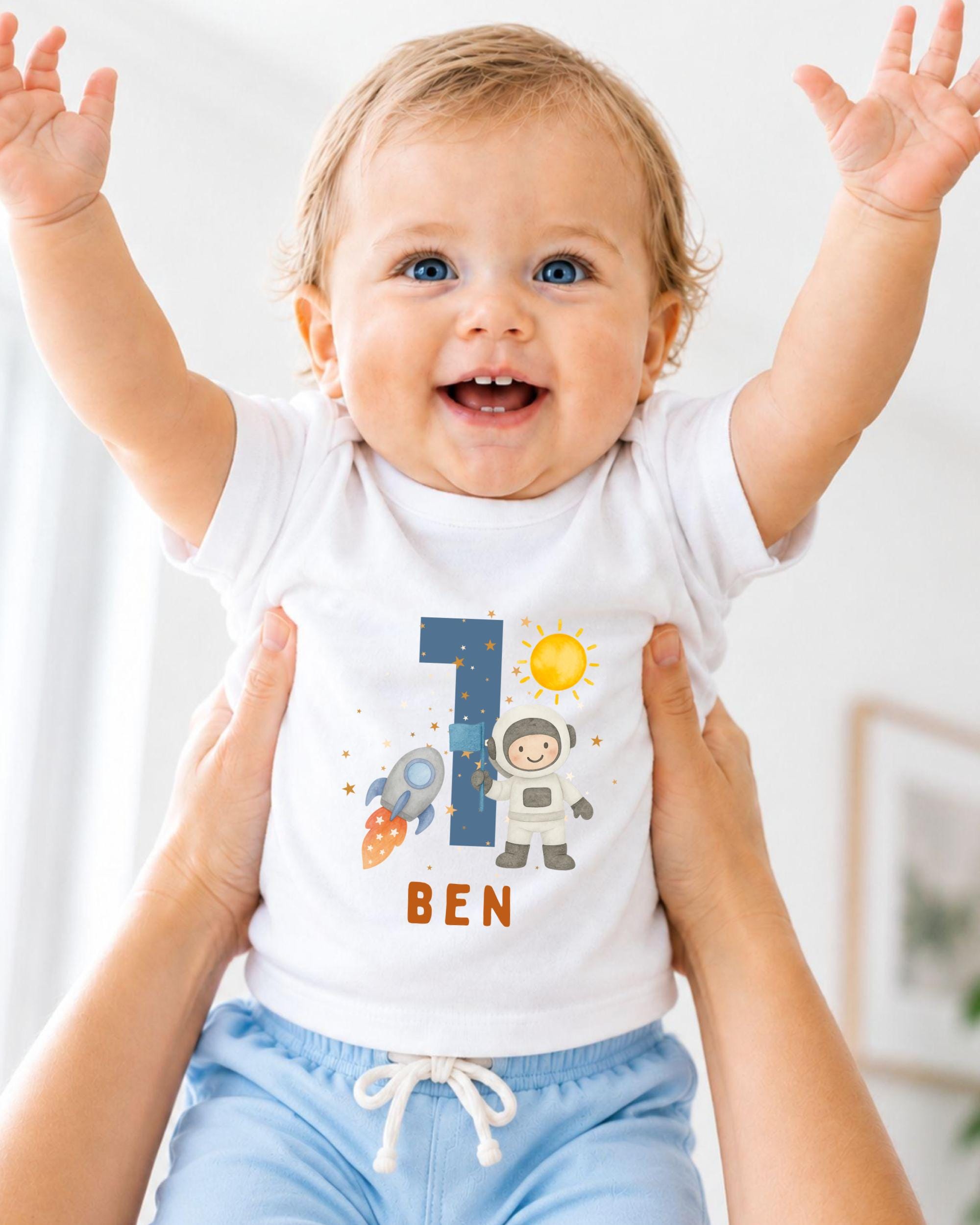 A smiling baby wearing a white t-shirt with a space-themed design and the number 1, being held up by someone's arms.