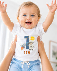 A smiling baby wearing a white t-shirt with a space-themed design and the number 1, being held up by someone's arms.