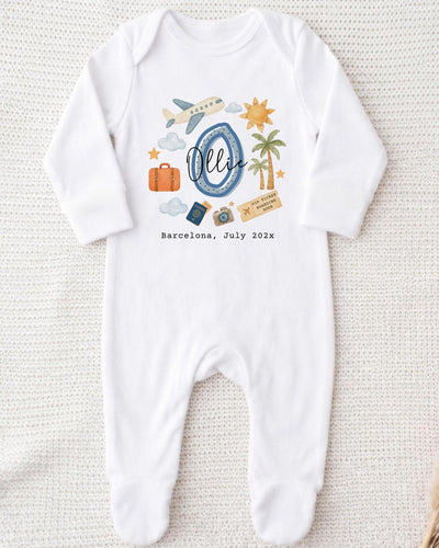A white onesie with a graphic design depicting various travel-related items such as a suitcase, airplane, palm tree, and a sun. The text "Barcelona, July 2020" is also printed on the onesie.