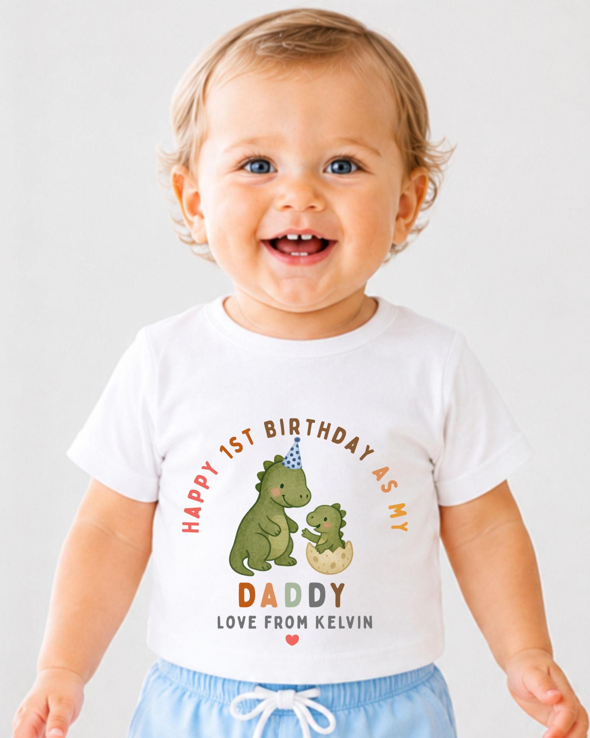 A smiling baby boy wearing a white t-shirt with a dinosaur and text that says "Happy 1st Birthday as my Daddy Love from Kevin".