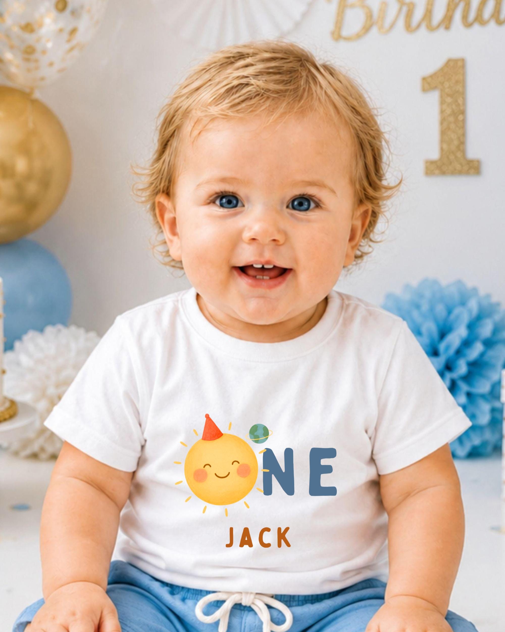 A smiling baby boy wearing a white t-shirt with a sun and planet design, sitting in front of a birthday party backdrop.