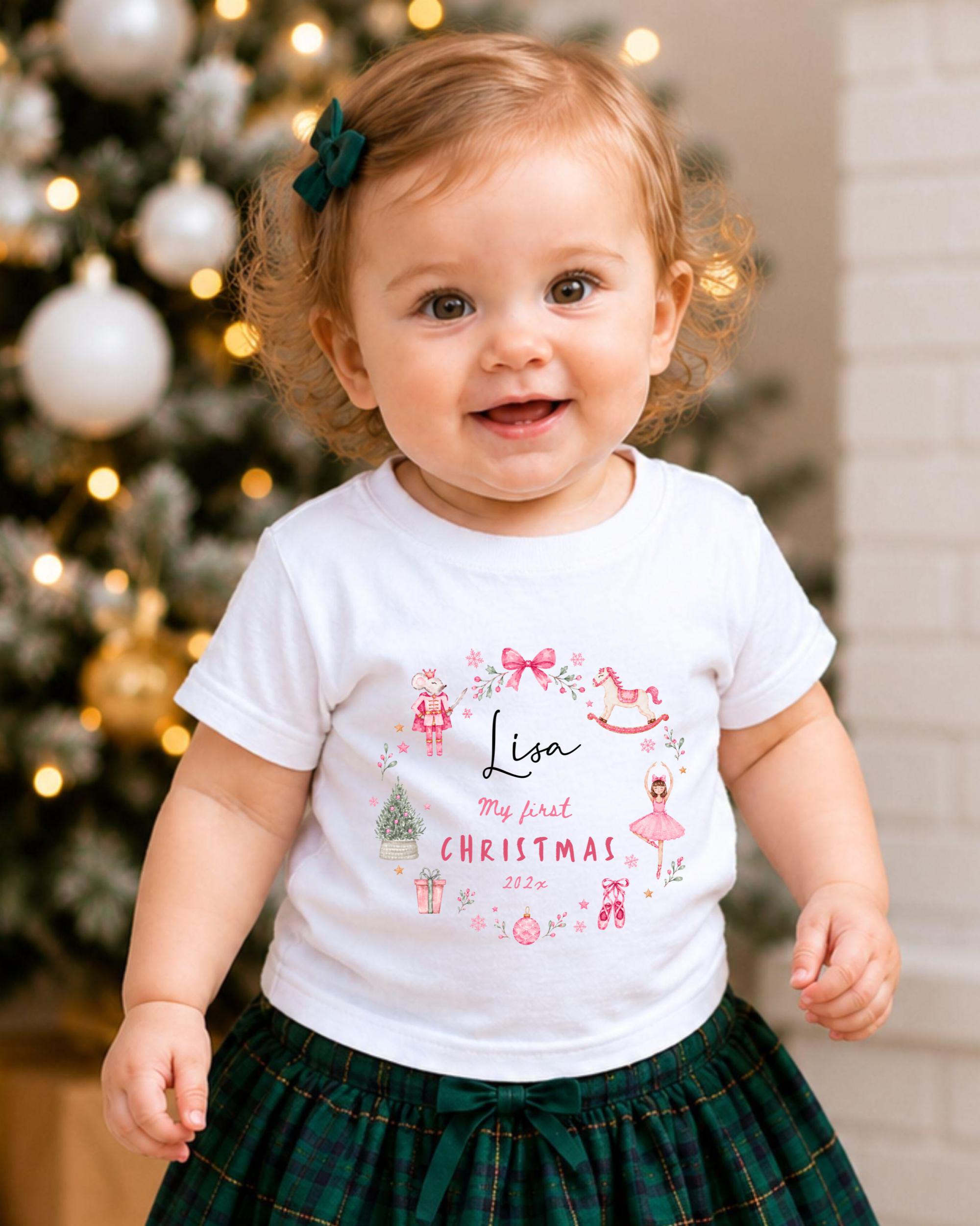 A smiling baby girl wearing a white t-shirt with a personalized design and text, standing in front of a Christmas tree.