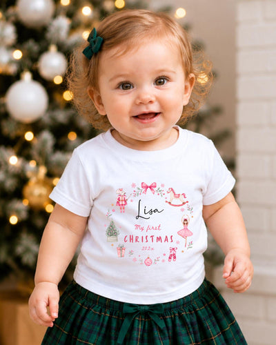 A smiling baby girl wearing a white t-shirt with a personalized design and text, standing in front of a Christmas tree.
