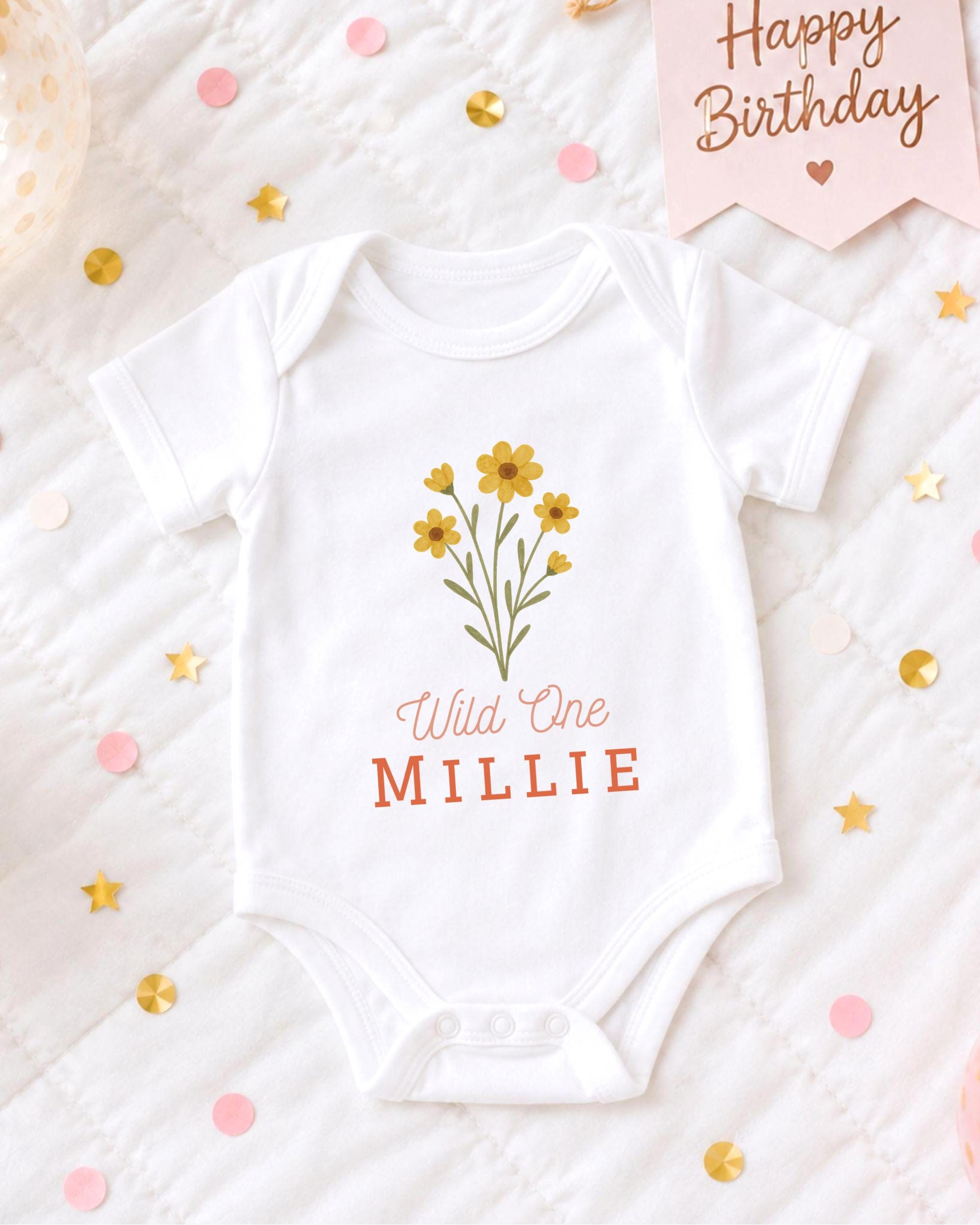 A white baby onesie with a flower design and the text "Wild One MILLIE" printed on it, placed on a bed with confetti and a birthday card.
