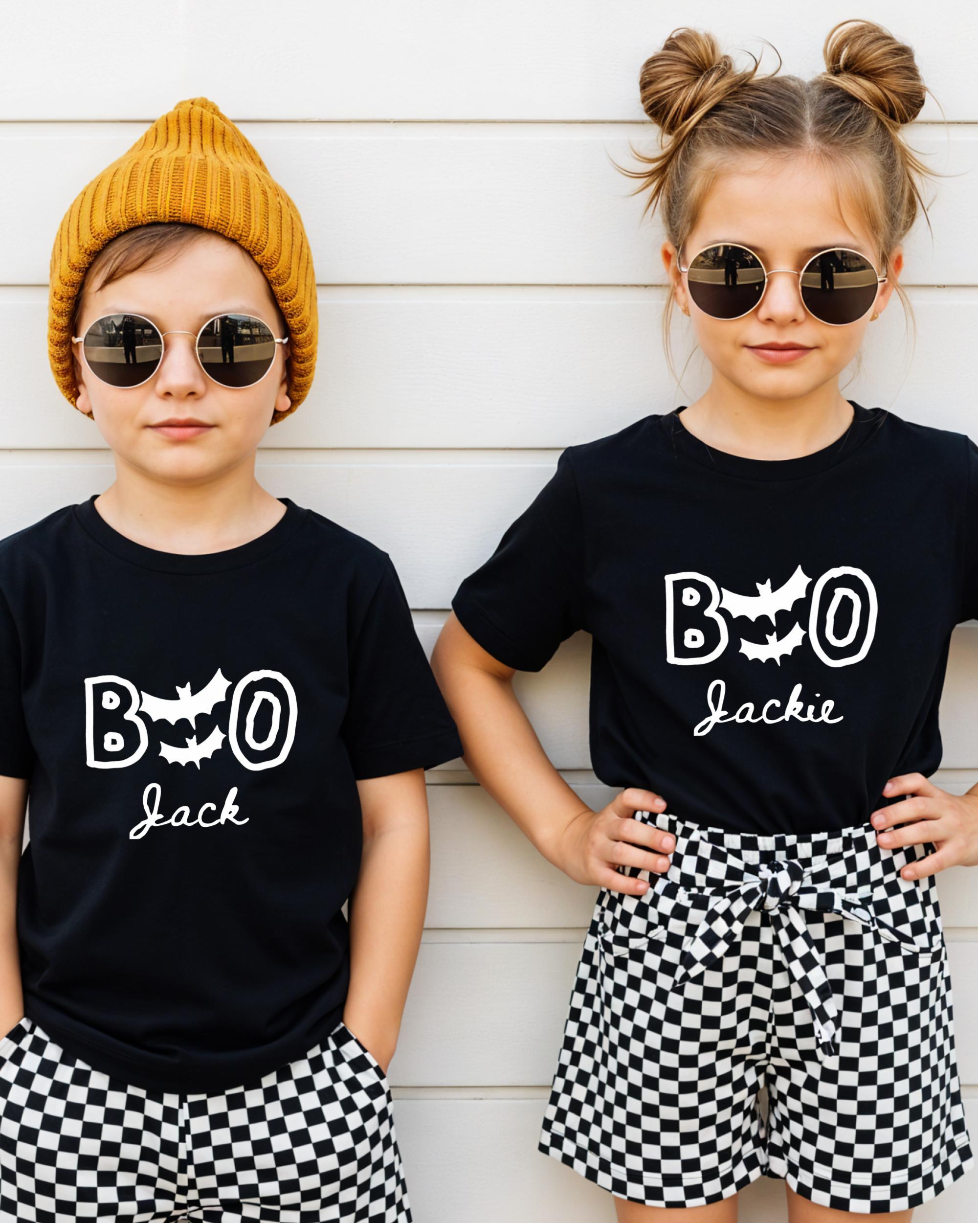 Two children wearing black t-shirts with the text "BOO Jackie" and round sunglasses, standing against a white wall.