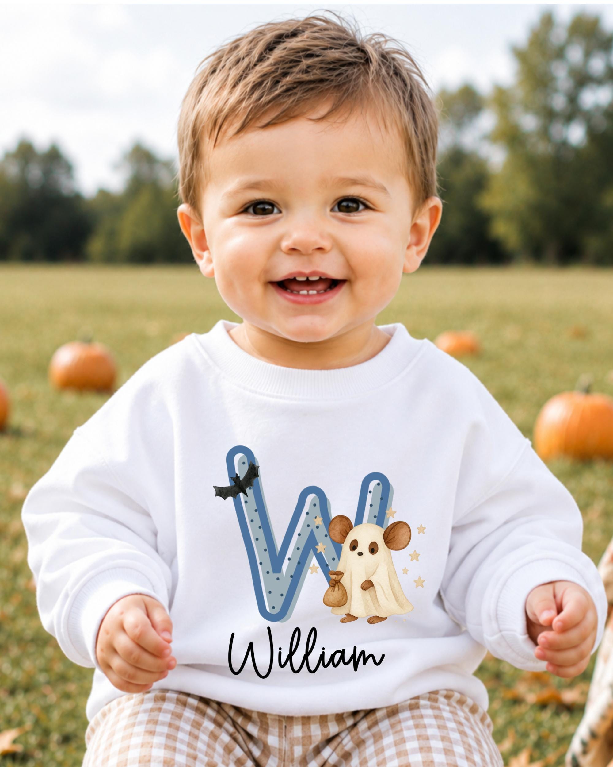A smiling baby boy wearing a white sweatshirt with a letter "W" and a ghost character, sitting in a field with pumpkins in the background.
