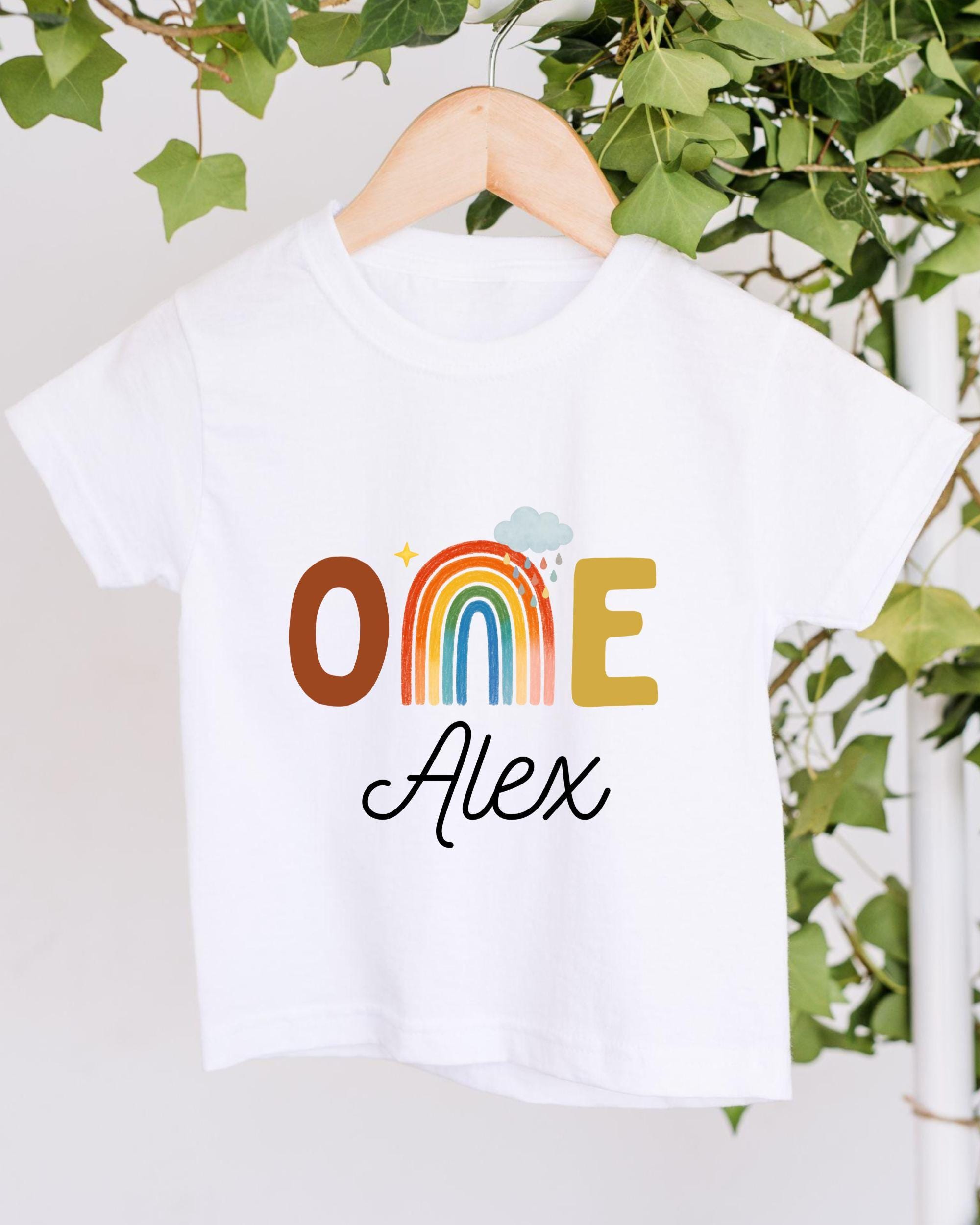 A white t-shirt with a colorful graphic design and the name "Alex" printed on it, hanging on a wooden hanger.