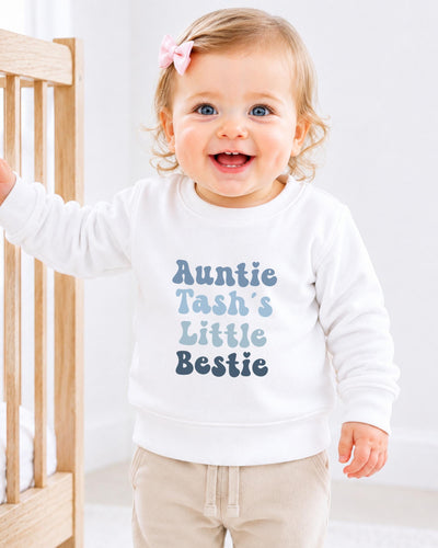 A smiling baby girl wearing a white sweatshirt with the text "Auntie Tash's Little Bestie" printed on it.