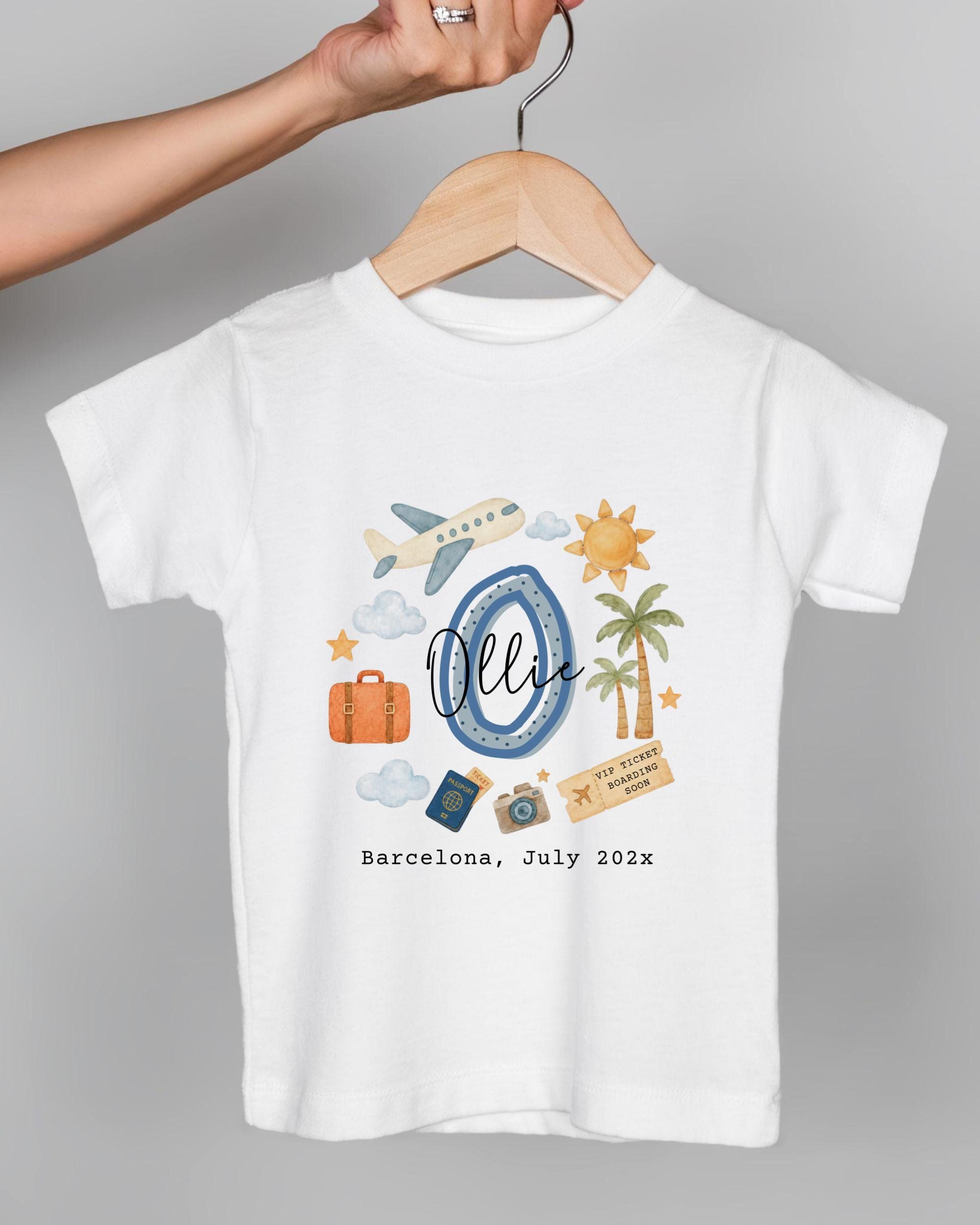 A white t-shirt with a graphic design featuring various travel-related icons, including a suitcase, a plane, a palm tree, and a sun. The text on the t-shirt says "Barcelona, July 2023".