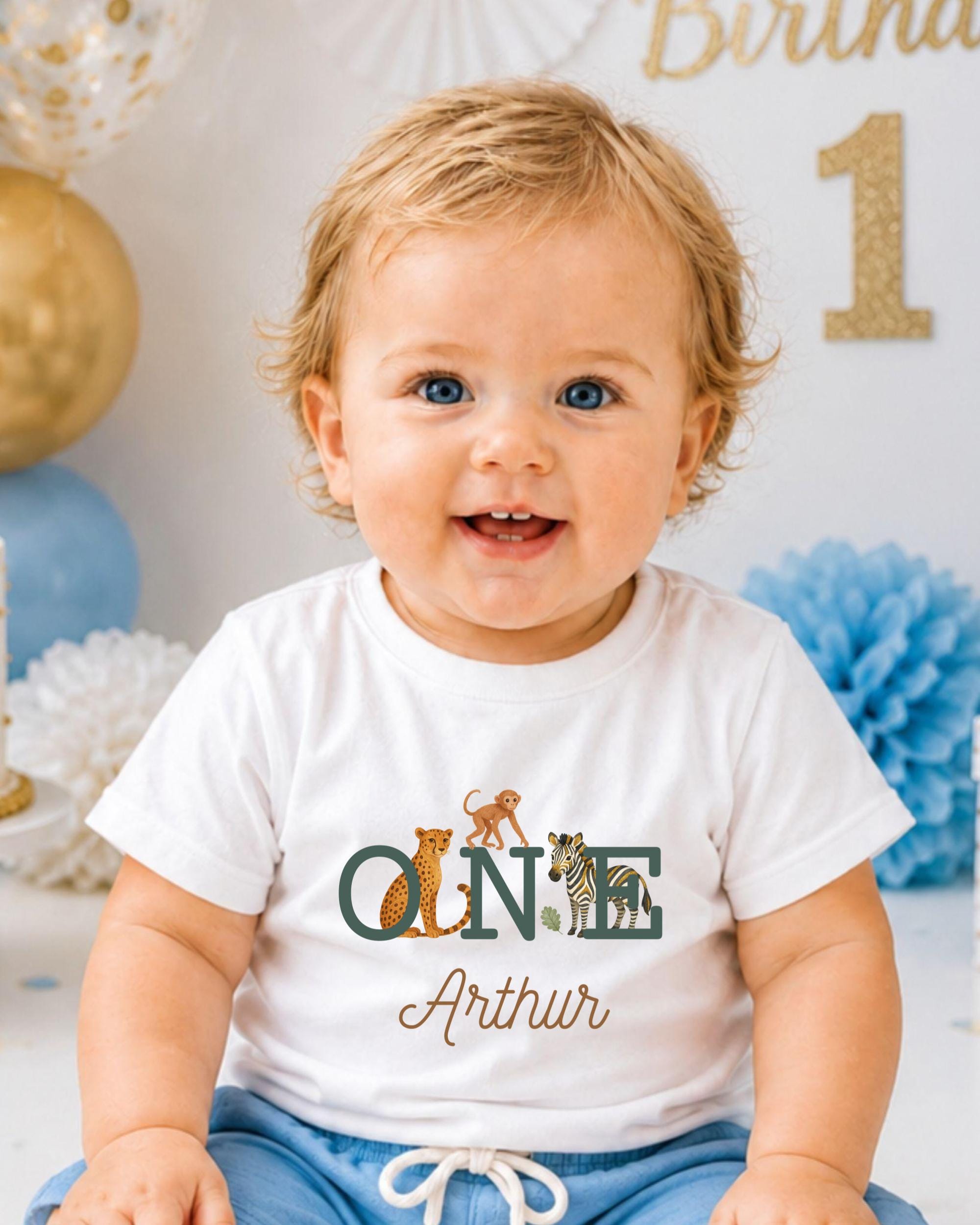 A smiling baby boy wearing a white t-shirt with a personalized name and animal print, sitting in front of a birthday party backdrop with balloons and confetti.