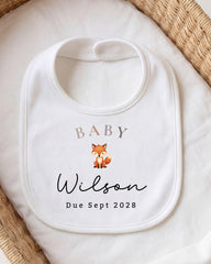A white baby bib with a personalized name and date printed on it, placed on a white cloth.
