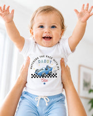 A smiling baby wearing a white t-shirt with a racing car graphic and the text "WATCHING THE RACE WITH MY DADY" is being held up by someone's arms.