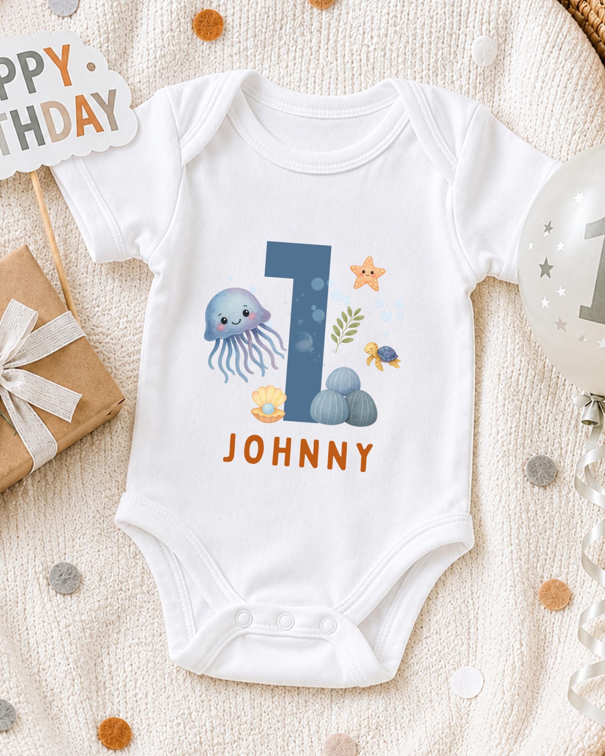 A white onesie with a blue number 1 and various marine life designs, including an octopus, starfish, and sea turtle, is displayed on a white blanket.