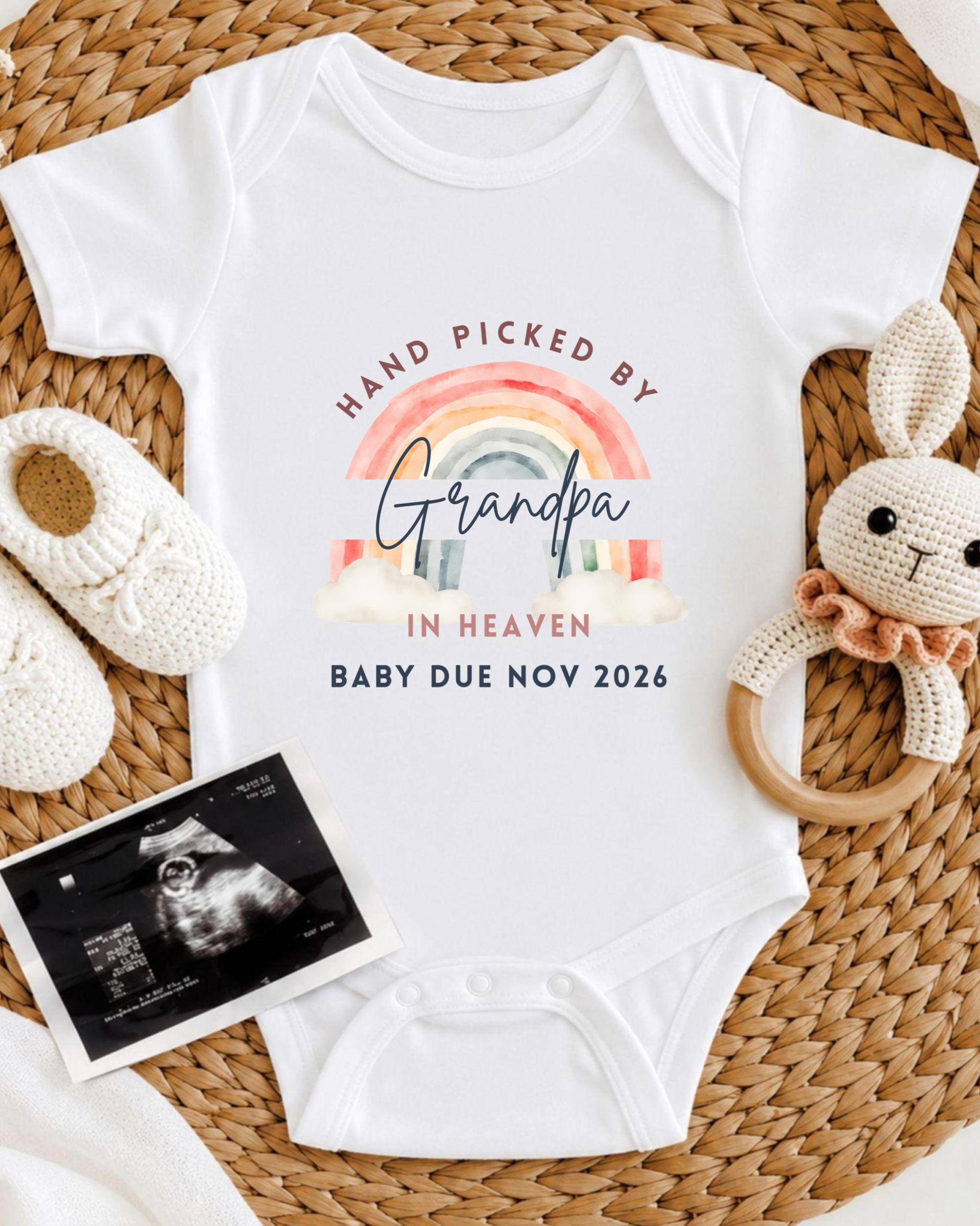 A white baby onesie with a rainbow graphic and text, along with a pair of white crocheted shoes and a baby rattle, all arranged on a woven mat.