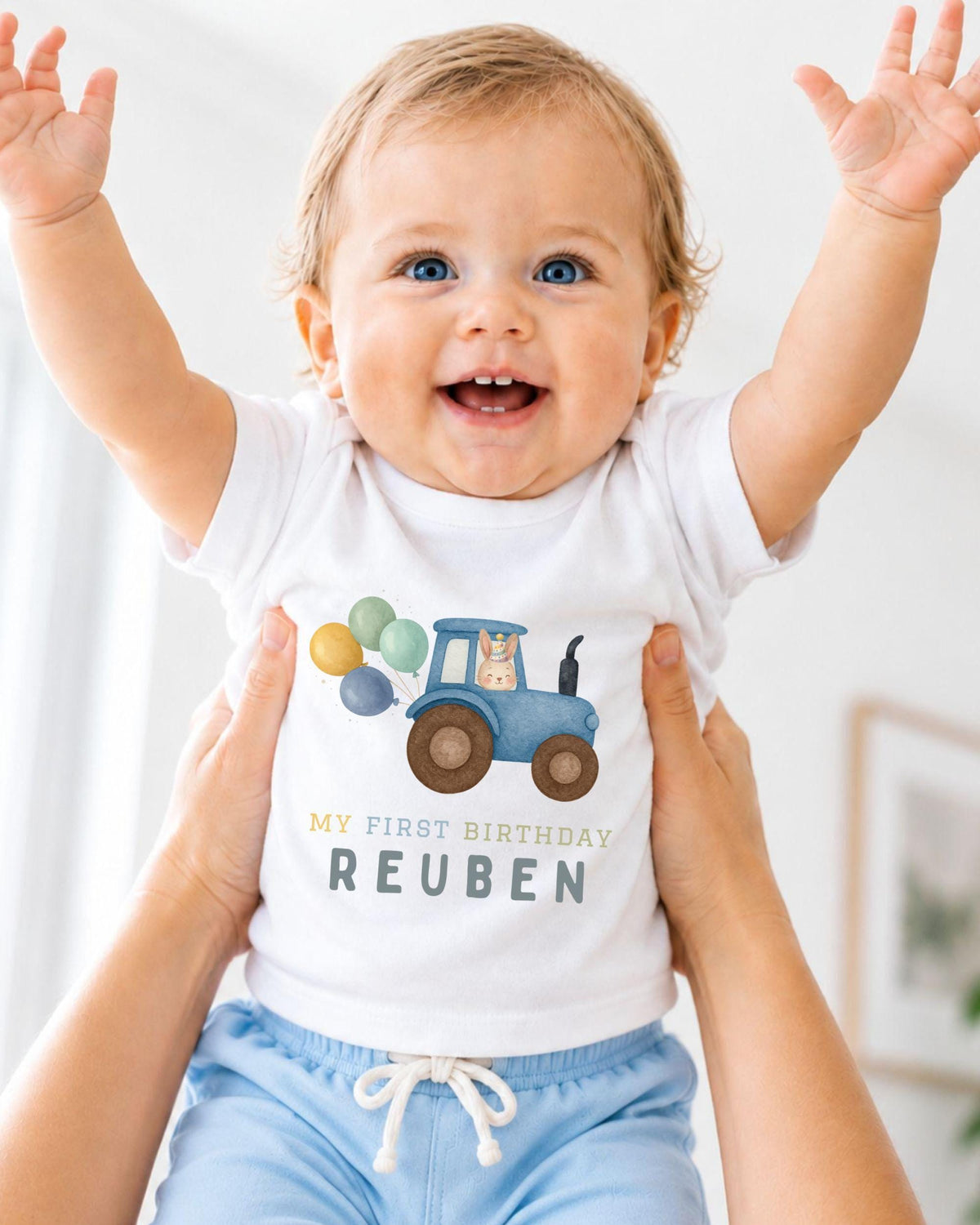 A smiling baby wearing a white t-shirt with a tractor graphic and the text "My first birthday Reuben" is being held up by someone's arms.