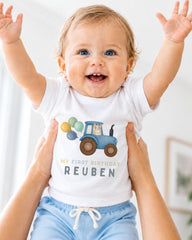 A smiling baby wearing a white t-shirt with a tractor graphic and the text "My first birthday Reuben" is being held up by someone's arms.